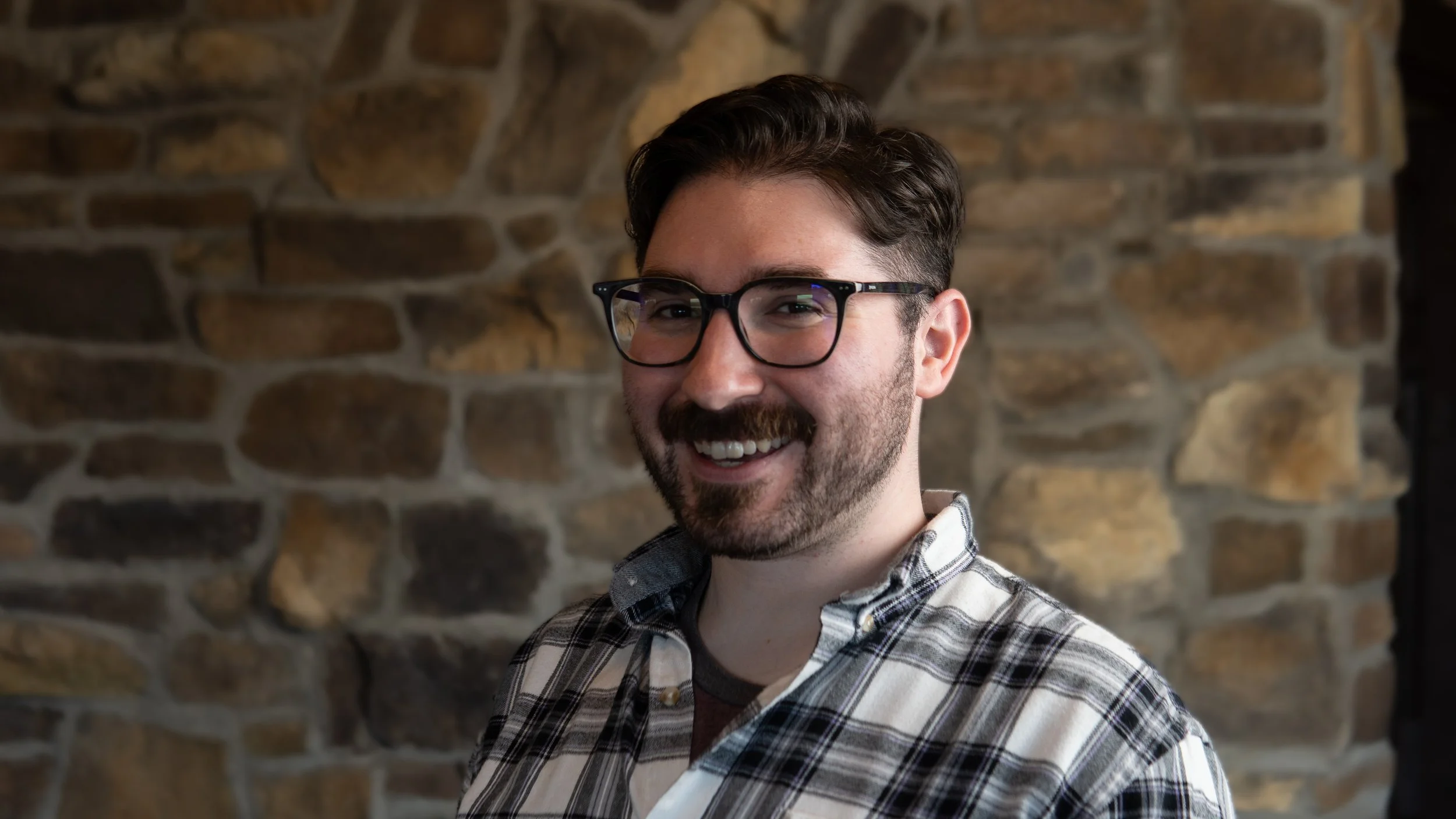 A smiling man with dark hair, glasses, and a beard wearing a plaid shirt in front of a stone wall.