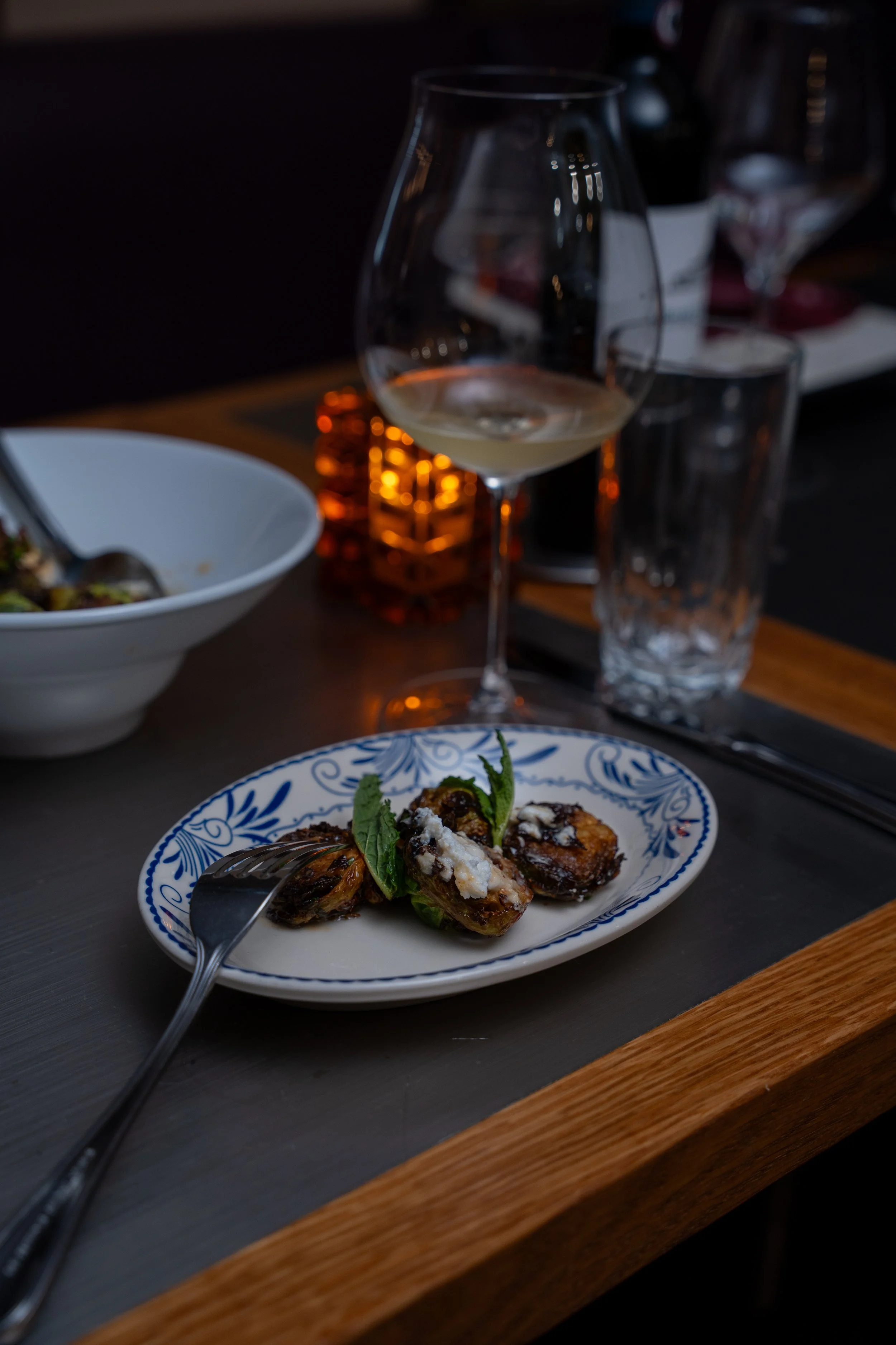 A white plate with blue decorative patterns contains small fried or roasted garnished bites, topped with cheese and fresh herbs, placed on a gray table with a fork. In the background, a wine glass with white wine, a tall empty glass, a candle, and bl