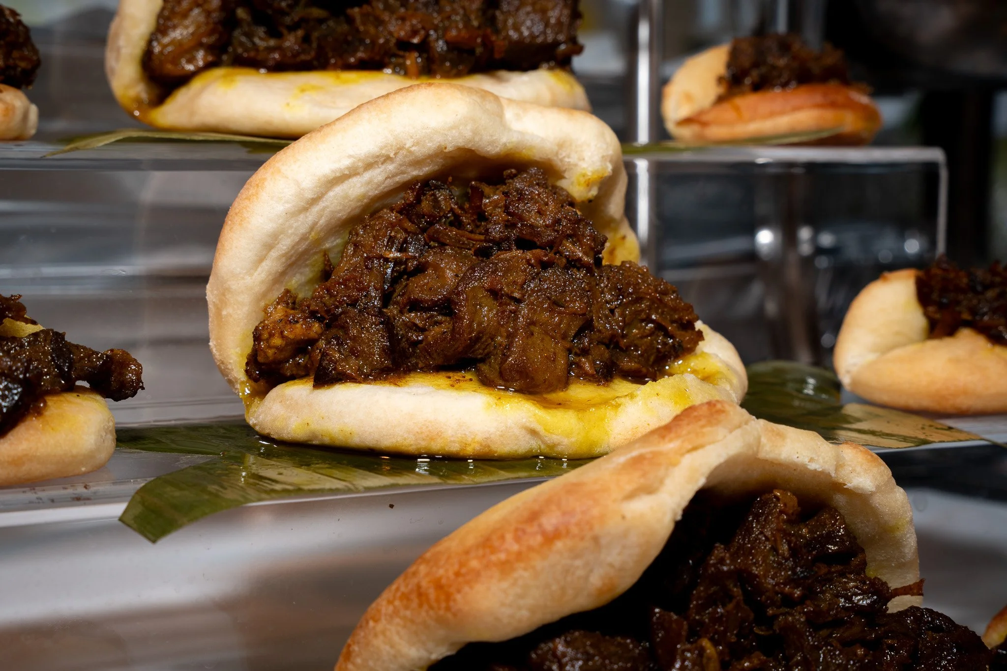Close-up of a meat-filled sandwich or bao with a soft, white bun and dark, cooked beef or pork filling. Several similar sandwiches are arranged on a display stand in the background.