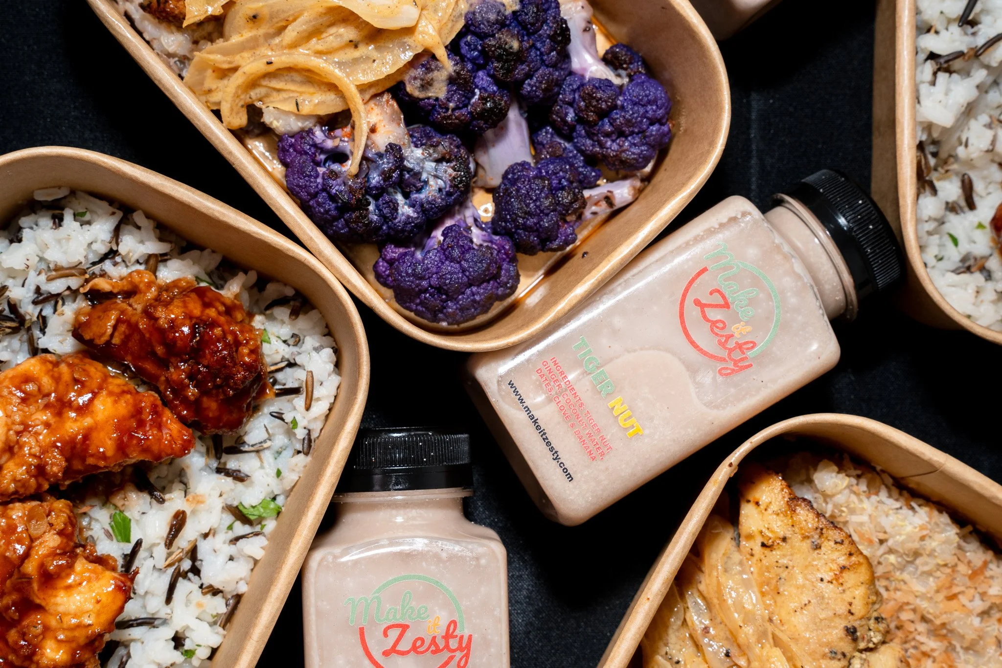 Close-up of various takeout food containers filled with rice, black cauliflower, spiralized vegetables, fried chicken, and dressings, arranged on a black surface.