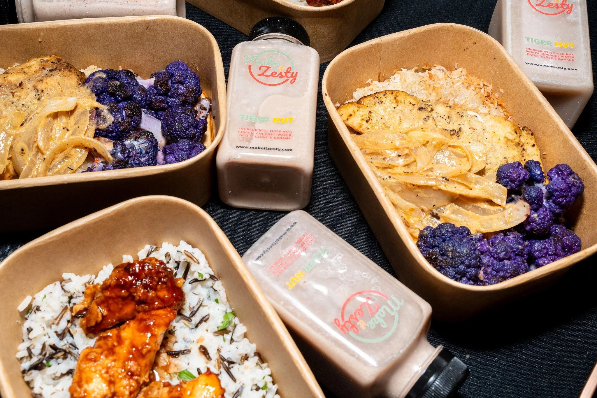 Bamboo bowls containing dishes like rice topped with sauce, roasted chicken, and purple cauliflower, with bottles of salad dressing labeled 'Make it Zesty' in the background.