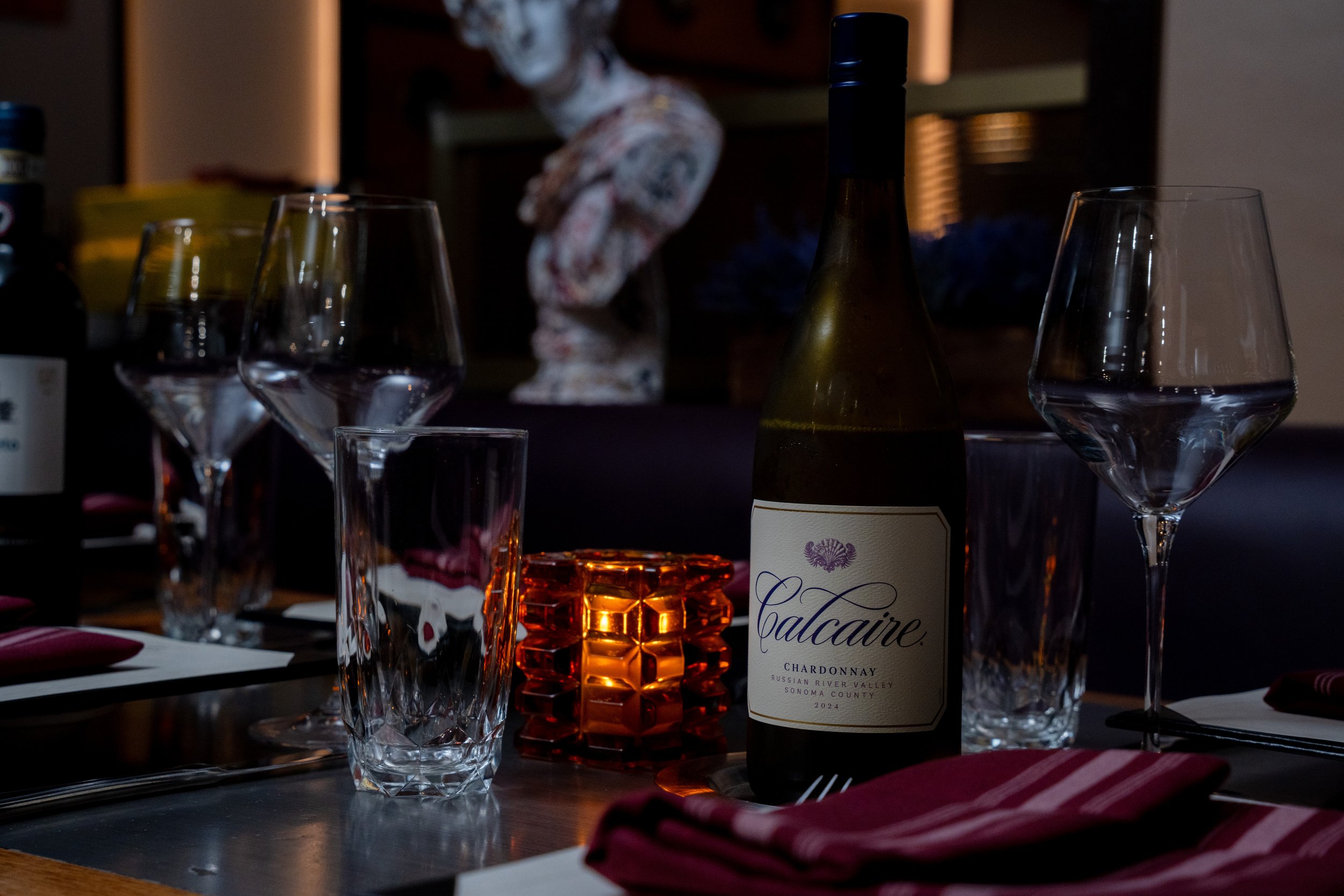 A table setting featuring a bottle of Calcaire Chardonnay wine, empty wine glasses, a small glass with a reddish drink, and a lit orange candle. In the background, a decorative skull and some other items are visible.