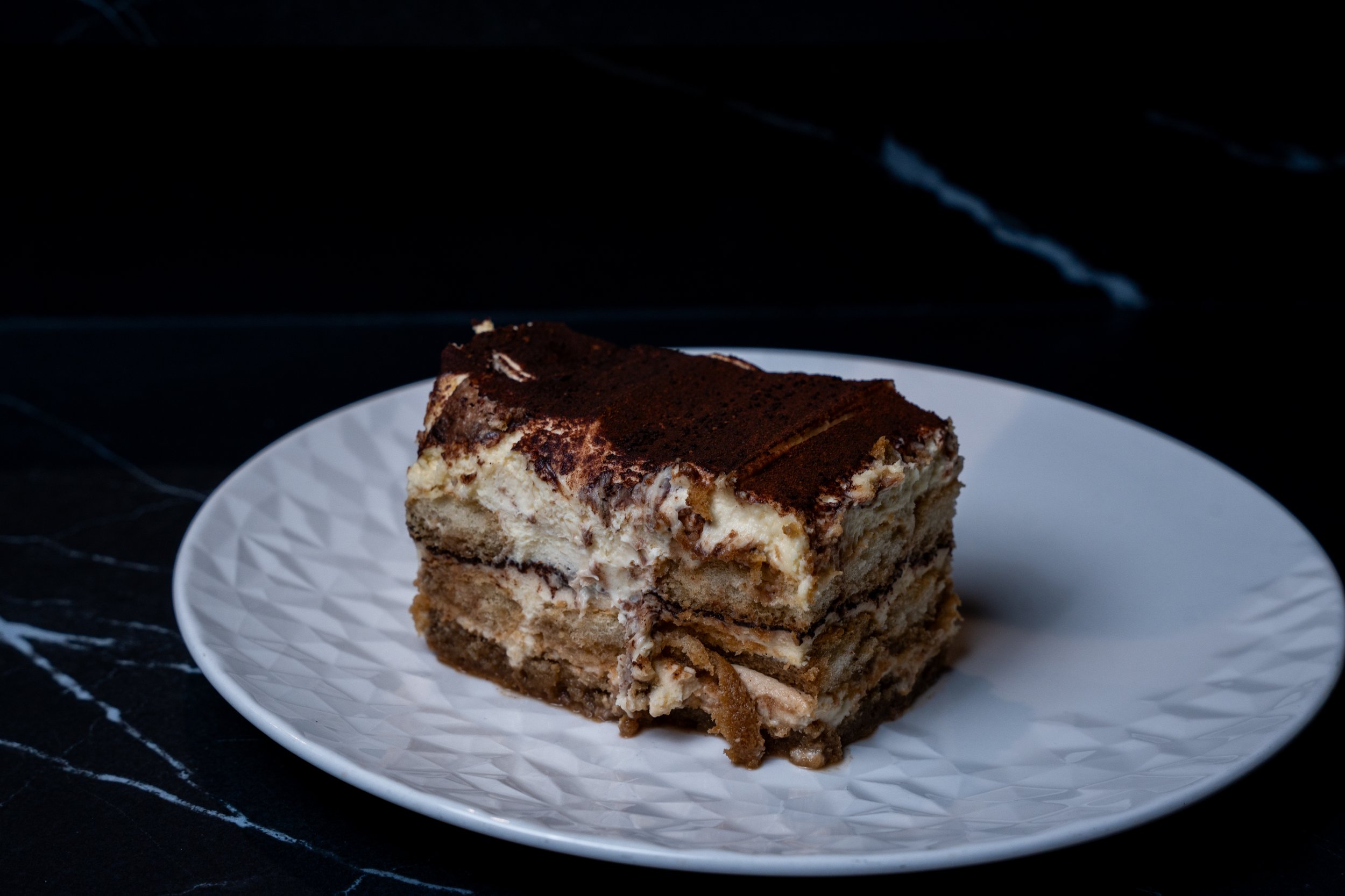 A slice of layered dessert with cream and chocolate on top, served on a white textured plate.