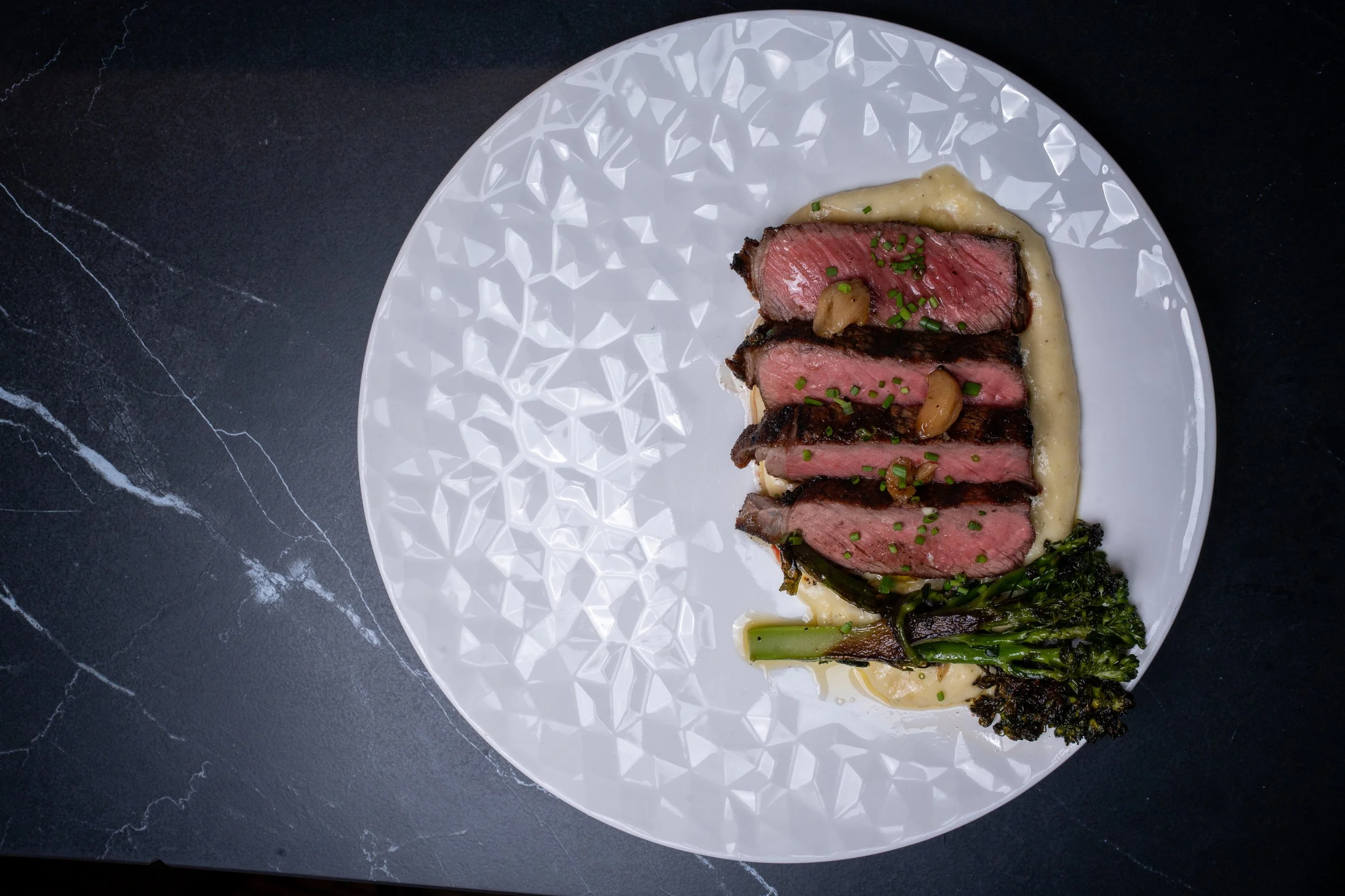 Slices of medium-rare steak with green herbs, garlic, and a creamy sauce on a decorative white plate, garnished with green chives and served with greens and roasted vegetables on a dark marble surface.
