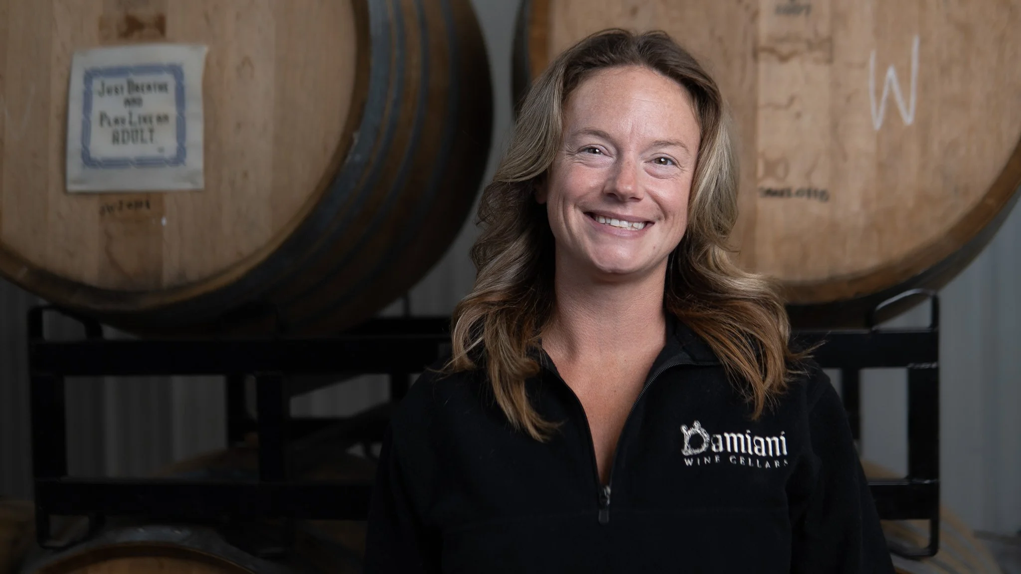 A smiling woman with light brown hair stands in front of wine barrels at Damiani Winery, wearing a black zip-up jacket with the winery's logo.