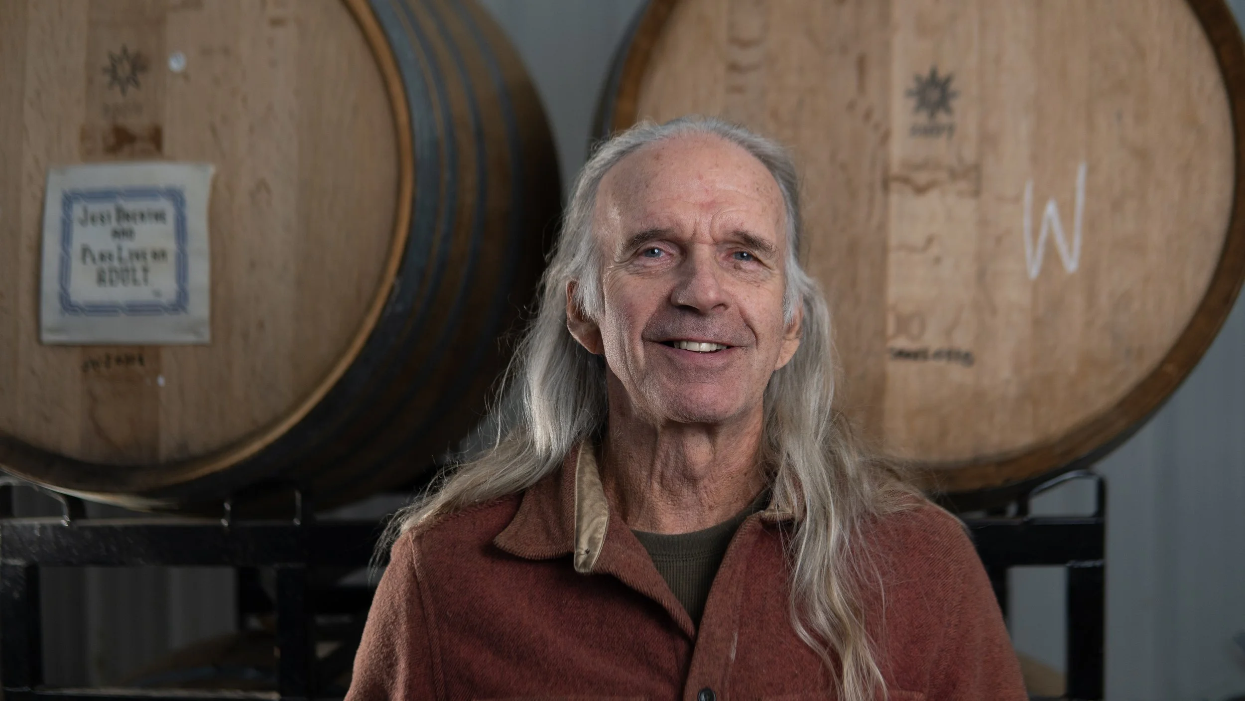 Older man with long gray hair smiling in front of large wooden barrels, one with a sign that says 'Just Barrels for Adults'.