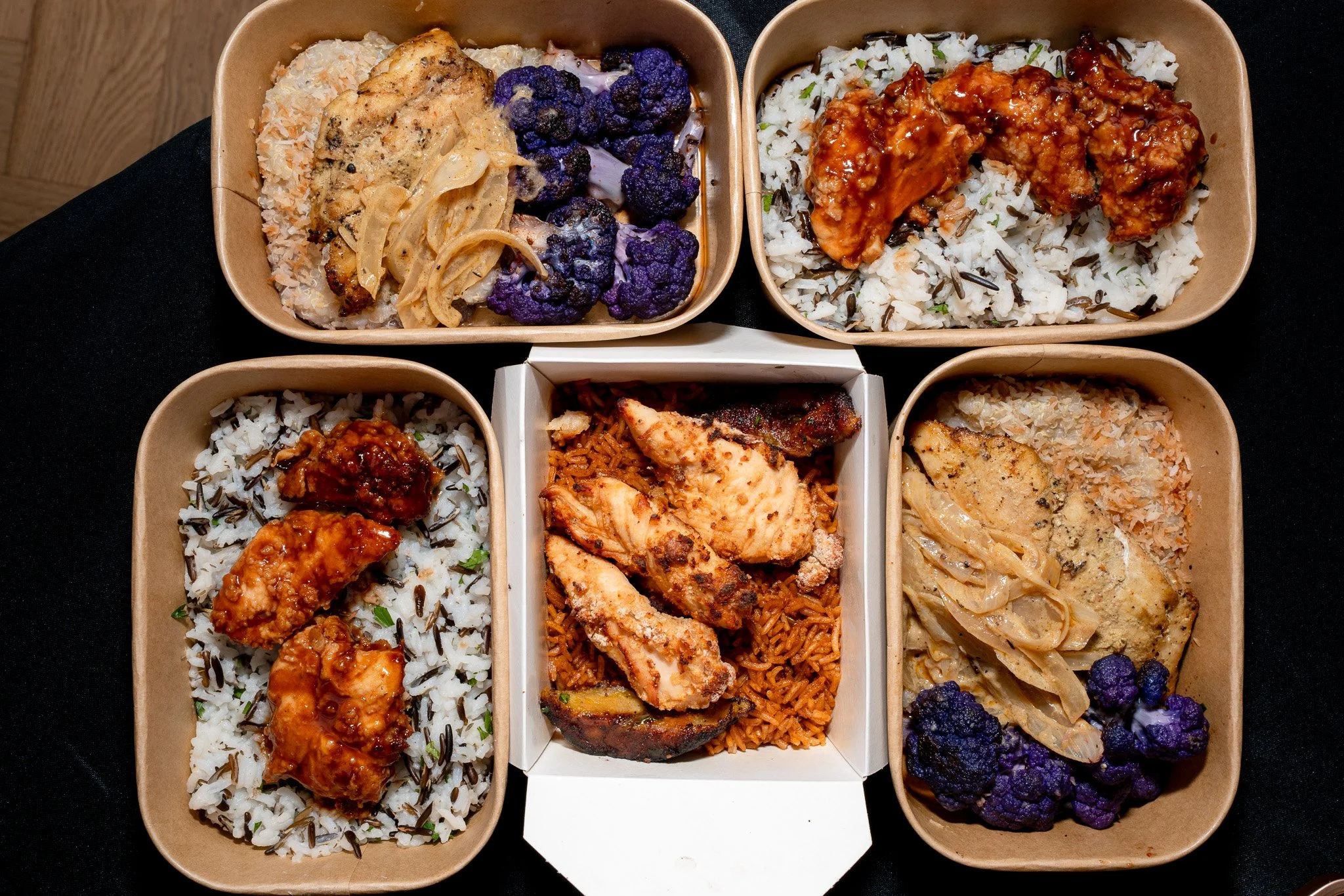Six rectangular sections of food in biodegradable containers, featuring rice with different toppings, a pasta dish with chicken, and roasted vegetables, arranged on a black surface.
