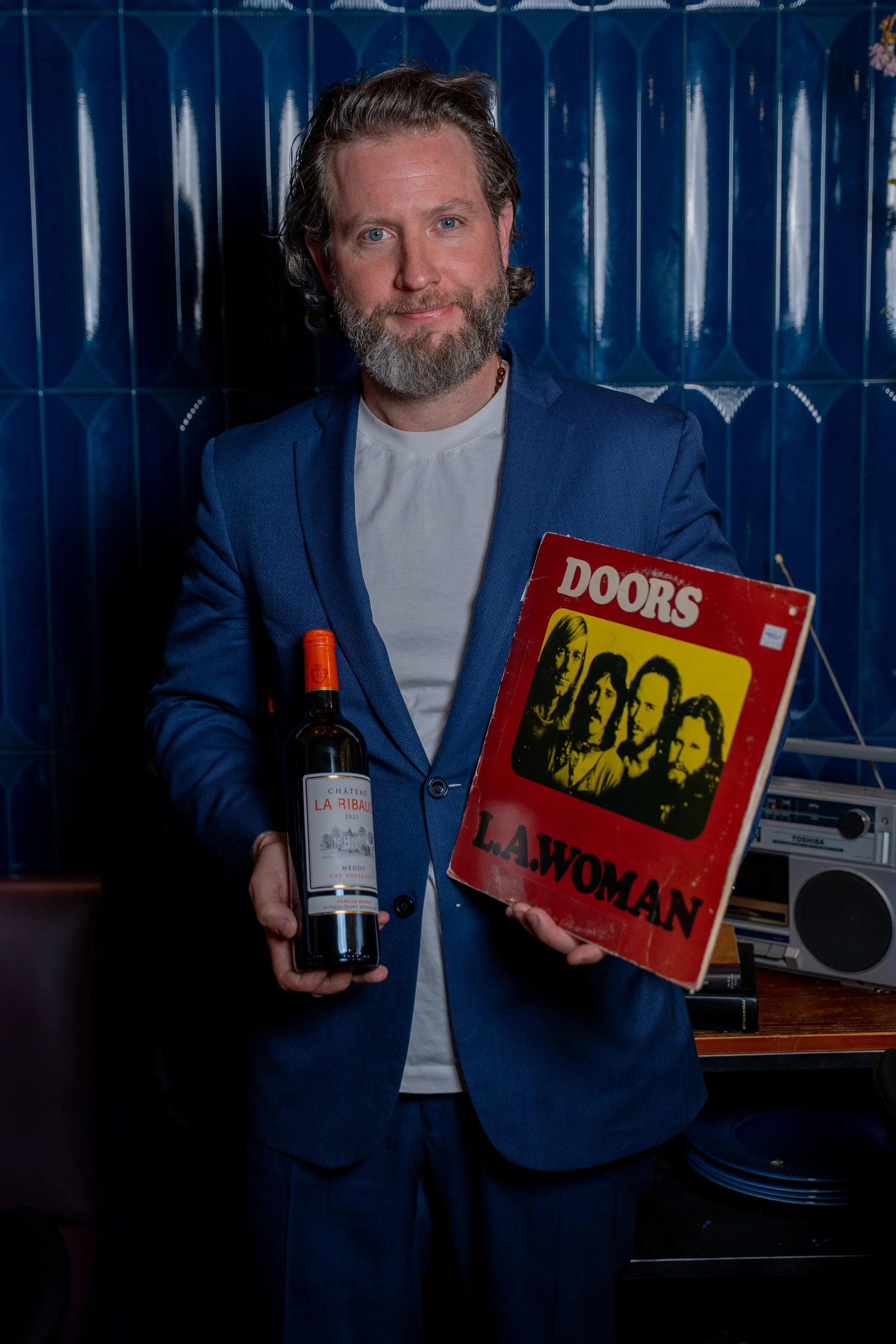 A man in a blue blazer with a beard and long hair, holding a bottle of wine labeled 'Château La Ribaude' in one hand and a vinyl record titled 'Doors' by L. A. Woman in the other hand. The background appears to be a tiled wall in a room with various 