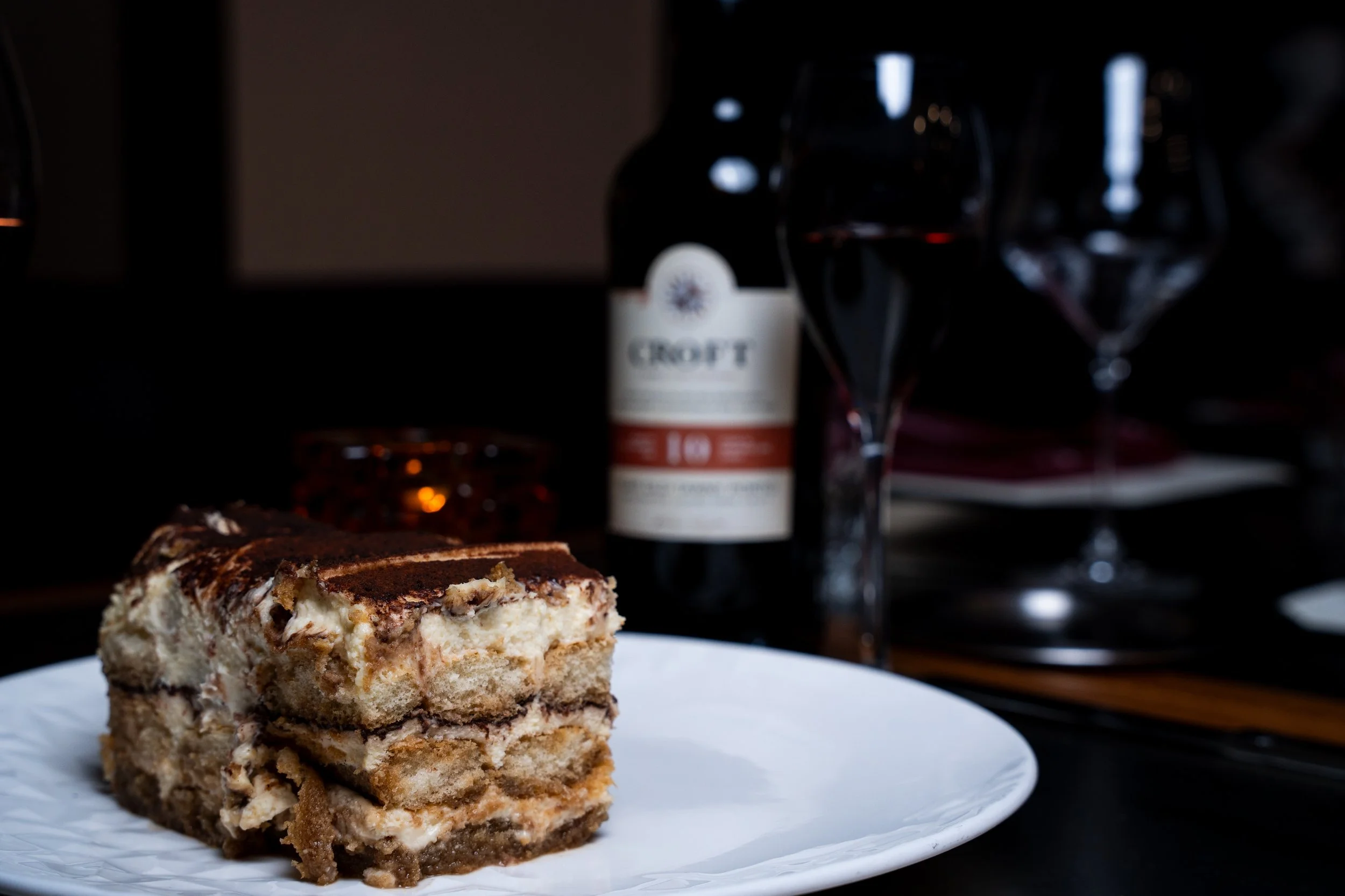 A plate with a slice of layered dessert, possibly tiramisu, with a dark background including a bottle of wine labeled Croft and two wine glasses.