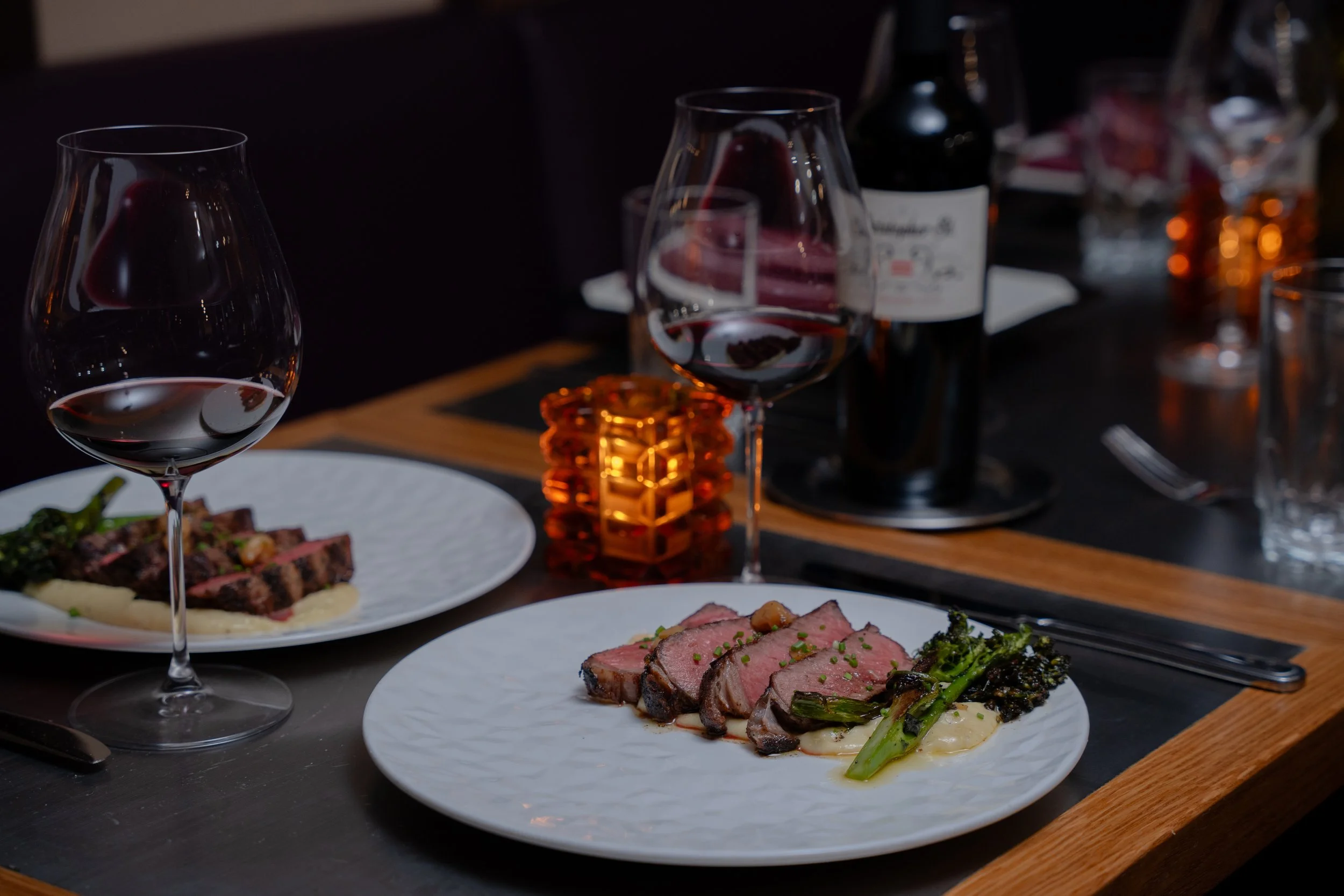 A dining table with plates of sliced meats, asparagus, and mashed potatoes, two glasses of red wine, a bottle of wine, and decorative candles, set in a dimly lit environment.