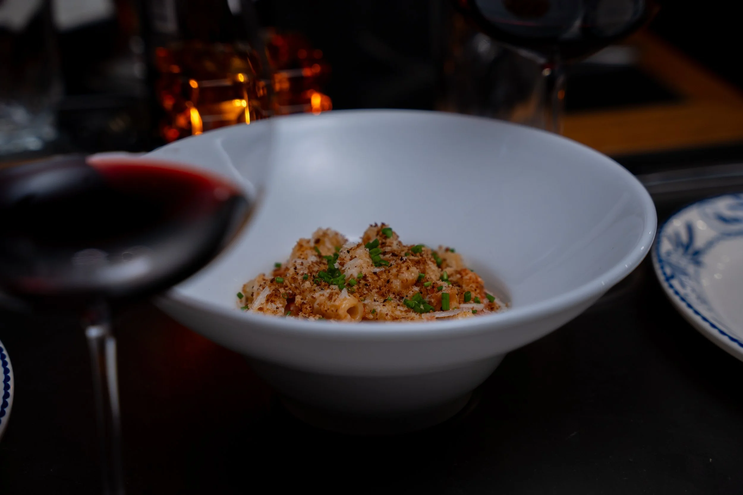 A white bowl of pasta topped with breadcrumbs and chopped green herbs, with red wine being poured into a glass nearby and a blue and white dish in the background.