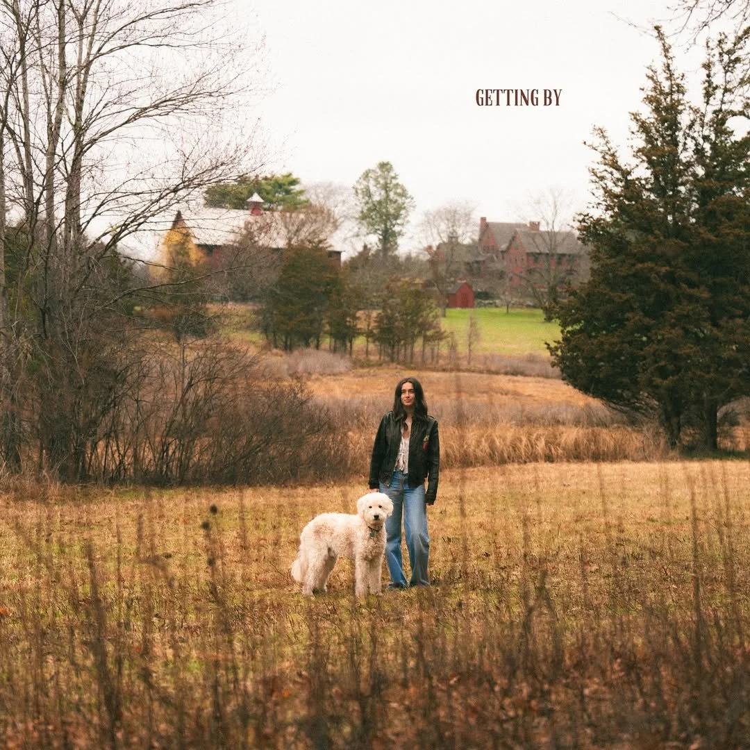 A woman standing in a field with a large white dog, trees, houses, and a cloudy sky in the background.