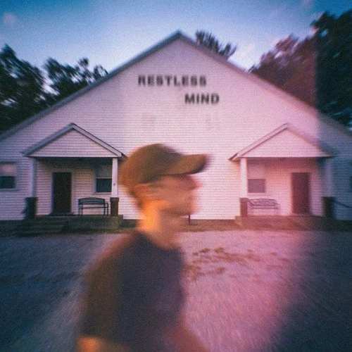 A blurry person walking in front of a white building with the words 'HUSTLE MIND' on it, and two small covered porches.