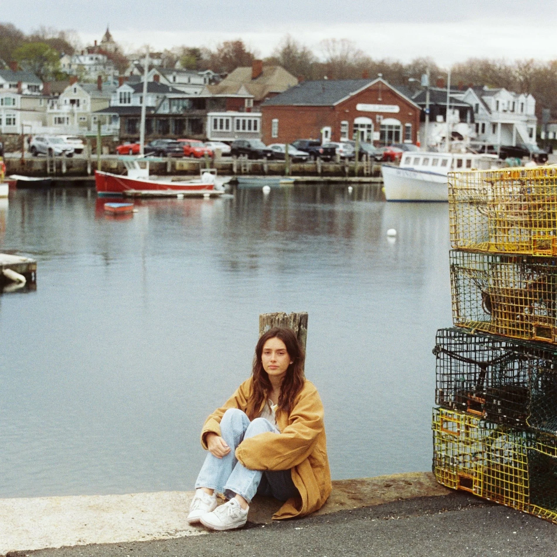 A young woman with long brown hair, wearing a tan jacket, white sneakers, and light blue jeans, is sitting on the edge of a dock by the water. Behind her, there are stacked yellow and black lobster traps and a harbor with boats and rows of houses along the shoreline.