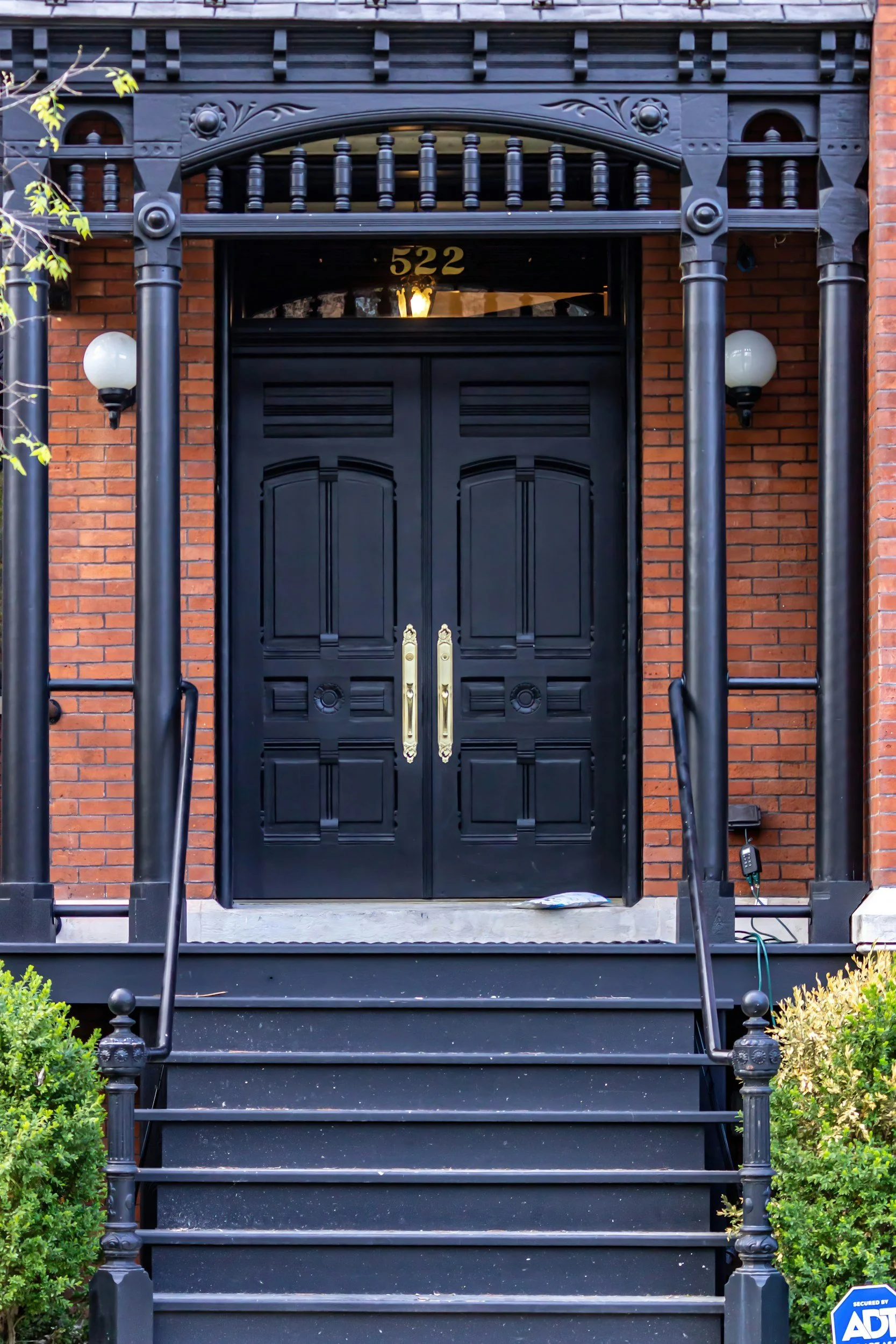 Black double doors with gold handles, set in a red brick building, accessed by black stairs with a metal railing, and flanked by wall-mounted lamps.