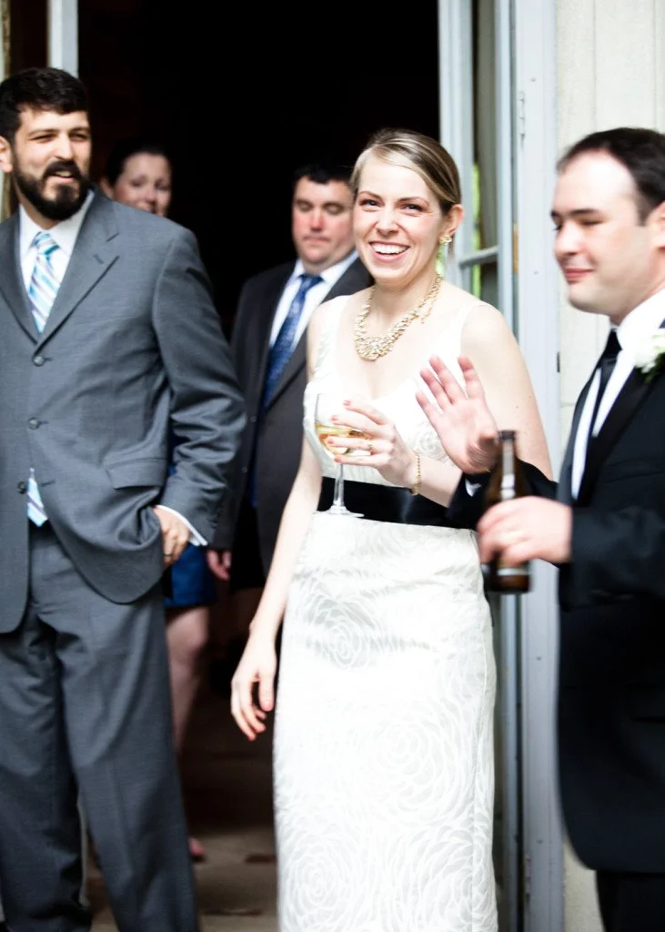 People dressed in formal attire at a social gathering, with a woman smiling and holding a glass of wine, while others around her are engaged in conversation.