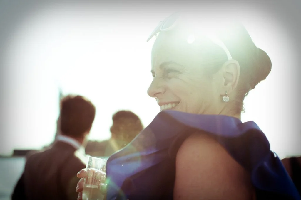 A woman in a blue dress smiling at a social gathering, with her hair styled in an updo and wearing earrings, holding a glass of clear beverage.