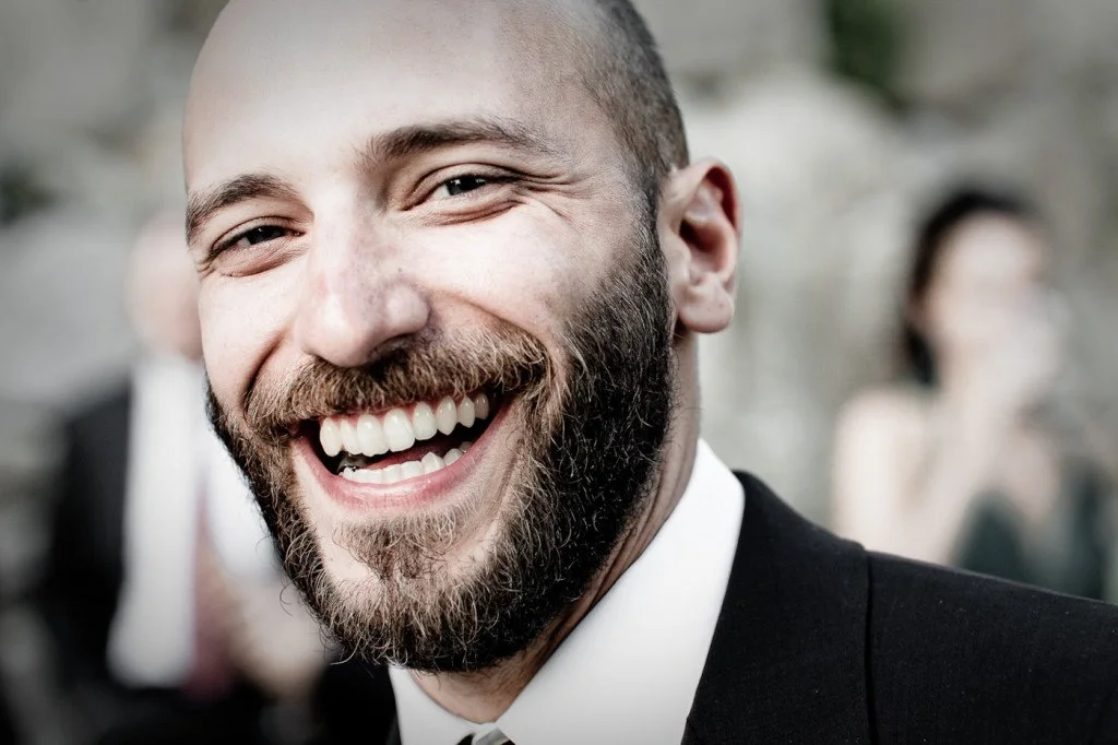 A man with a beard and short hair smiling in focus, blurred background with people dressed formally.
