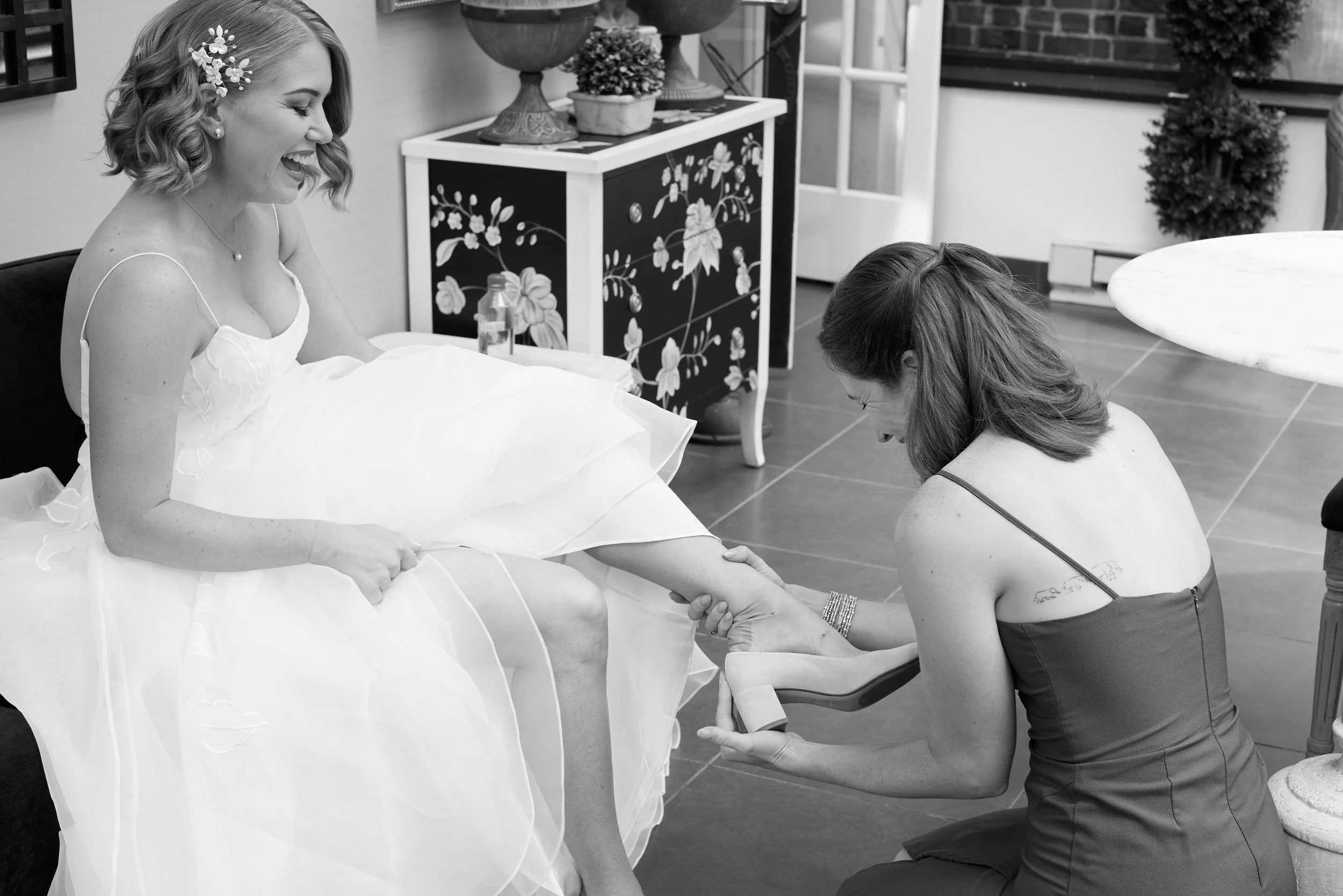 A woman in a white dress is sitting on a couch, smiling as another woman helps her put on a shoe.