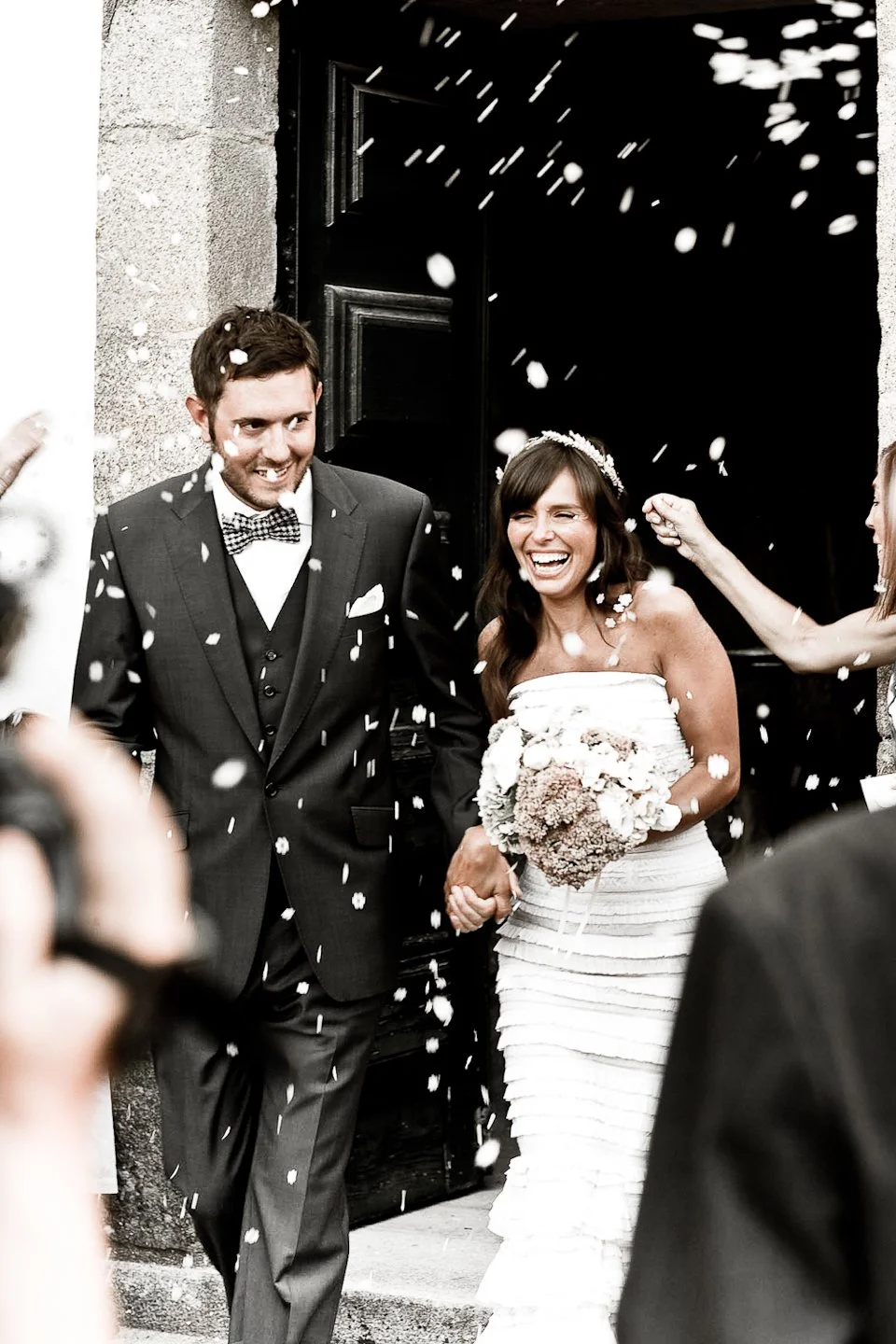 A newly married couple celebrating outside a church, smiling and holding hands, as confetti is thrown by wedding guests.