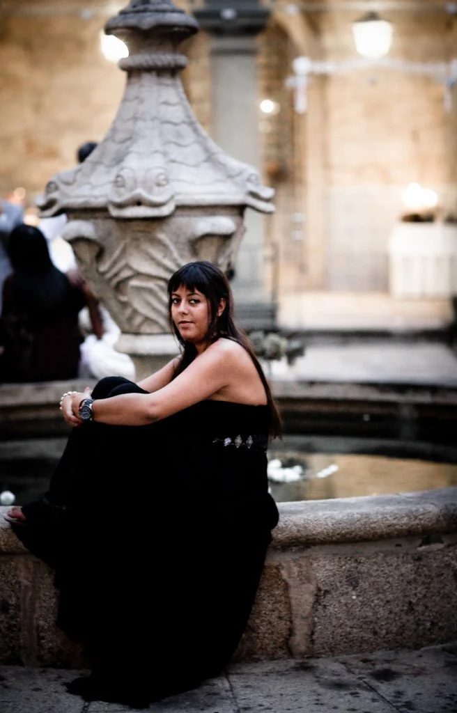 A woman with long dark hair and a black dress sitting on a stone fountain in a historic European courtyard.