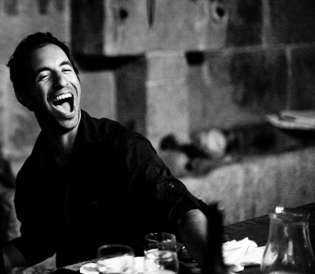 A man laughing and enjoying himself in a dimly lit setting, possibly a restaurant or bar, with glasses and a pitcher on the table.