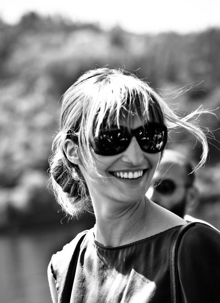 Black and white photo of a smiling woman wearing sunglasses outdoors, with wind-blown hair and blurred background.