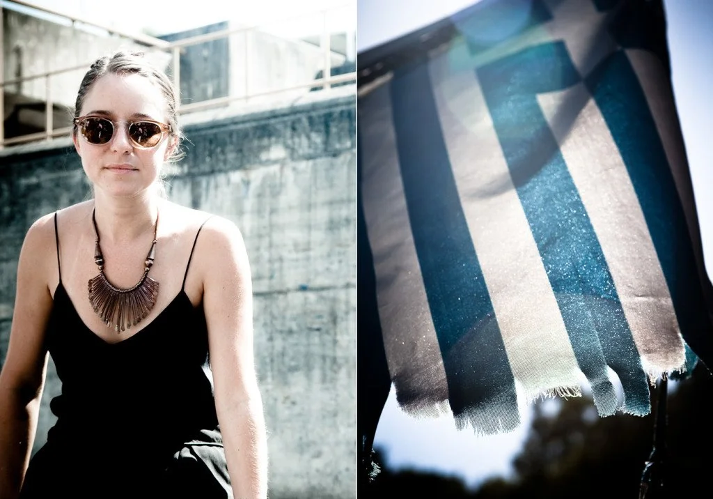 A woman wearing sunglasses, a black tank top, and a beaded necklace sitting outdoors near a concrete wall with a railing above, and a striped fabric hanging with sunlight reflecting off of it.
