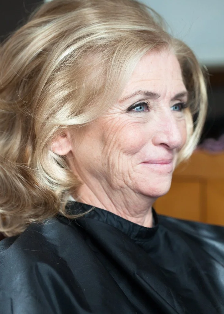 Close-up of a woman with blonde hair, blue eyes, and visible wrinkles, sitting in a black cape in a salon.