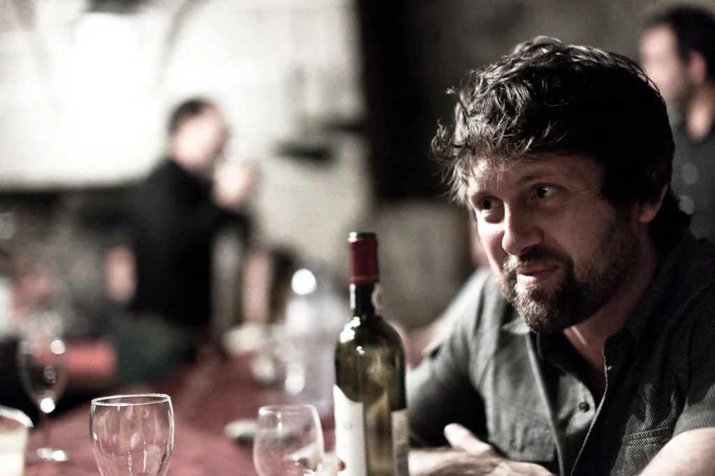 A man with curly hair and a beard sitting at a table in a dimly lit bar, with a bottle of wine and glasses in front of him, while other people are blurred in the background.