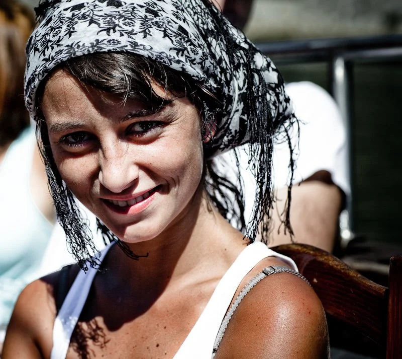 A woman with short dark hair wearing a black and white patterned headscarf, smiling outdoors in sunlight.