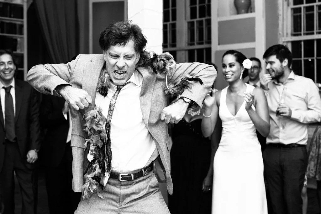 Man in a suit dancing enthusiastically at a party with several smiling guests around him, including a woman in a white dress and others in formal attire.