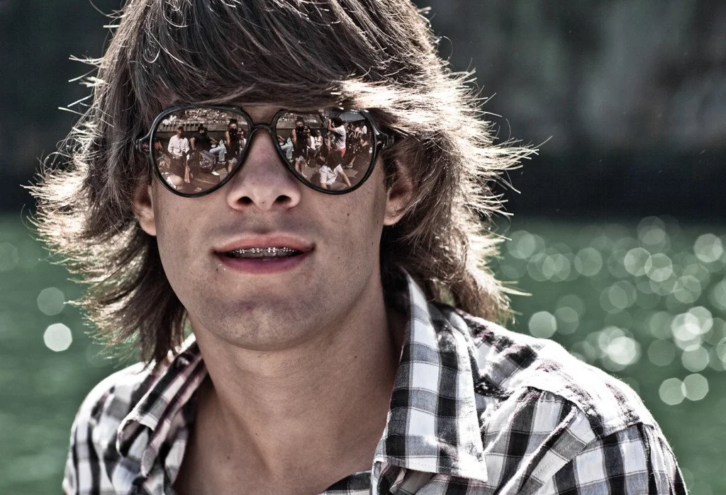 A young man with shoulder-length brown hair wearing sunglasses and a checkered shirt outdoors near water, with reflections visible in his sunglasses.