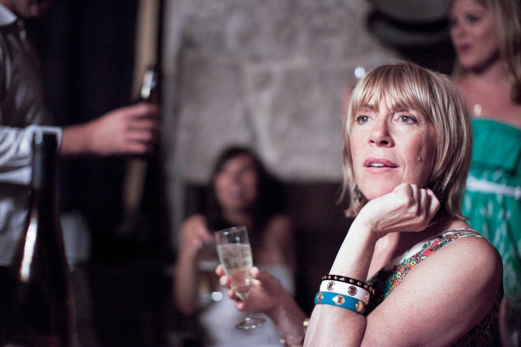 A woman with short blonde hair sitting at a social gathering, holding a glass of wine, with other people in the background.