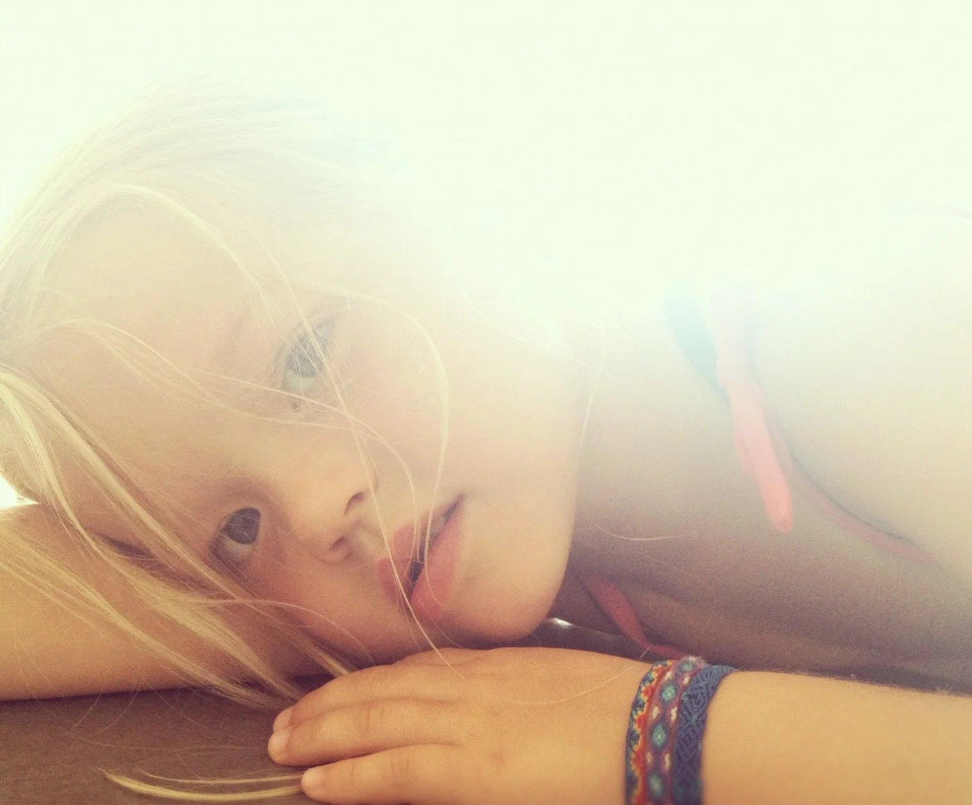 A young woman with blond hair resting her head on her arm, looking into the camera with a soft expression, wearing colorful bracelets on her wrist.