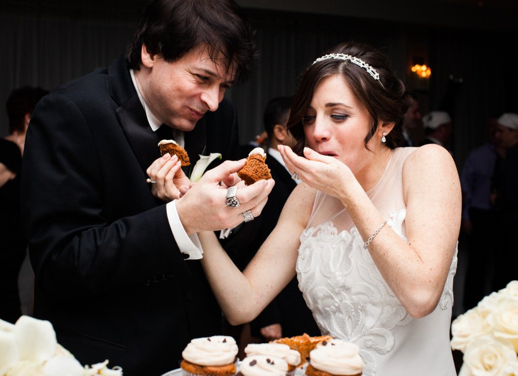 Bride and groom at a wedding cutting and sharing a cake, with cupcakes in the foreground and guests in the background.