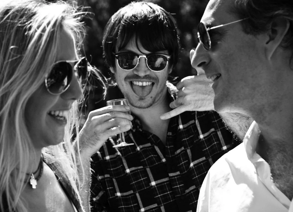 Three friends enjoying a sunny outdoor gathering, all wearing sunglasses. One woman is smiling, the man in the middle has a playful expression, and the man on the right is smiling and holding a glass of drink.