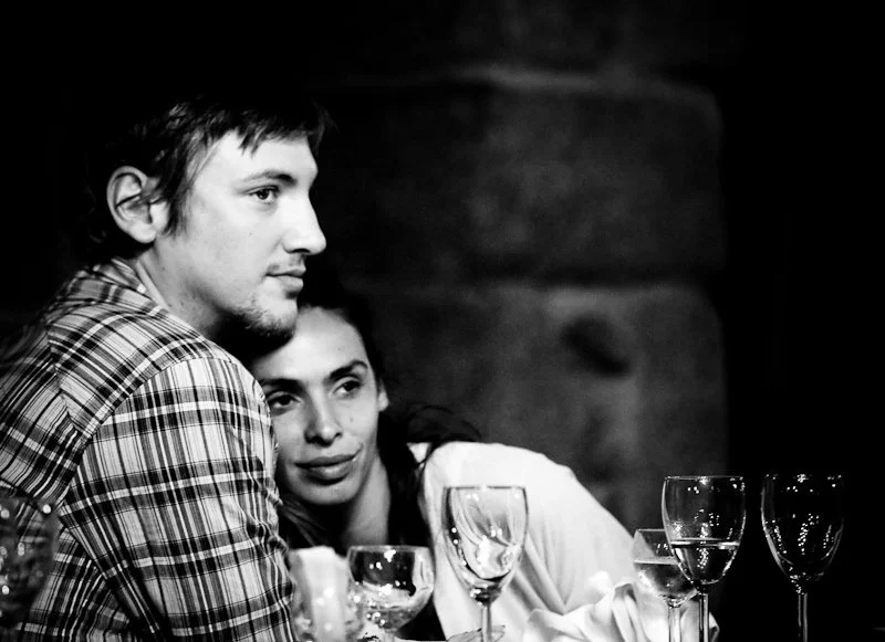 A black and white photo of a man and woman sitting at a table with empty wine glasses, with a dark background.