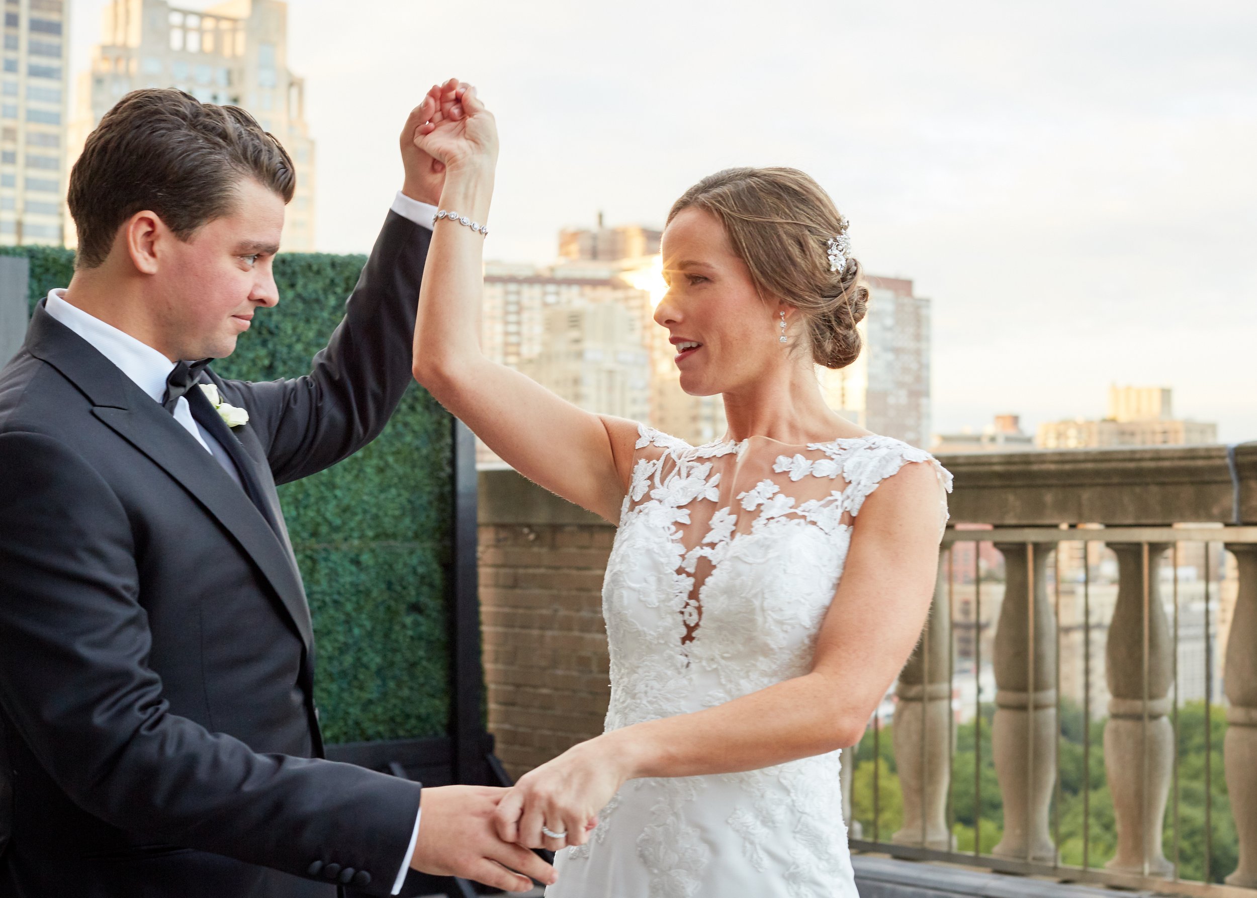 A bride in a white lace wedding dress and a groom in a black tuxedo dance on a balcony with city buildings in the background.