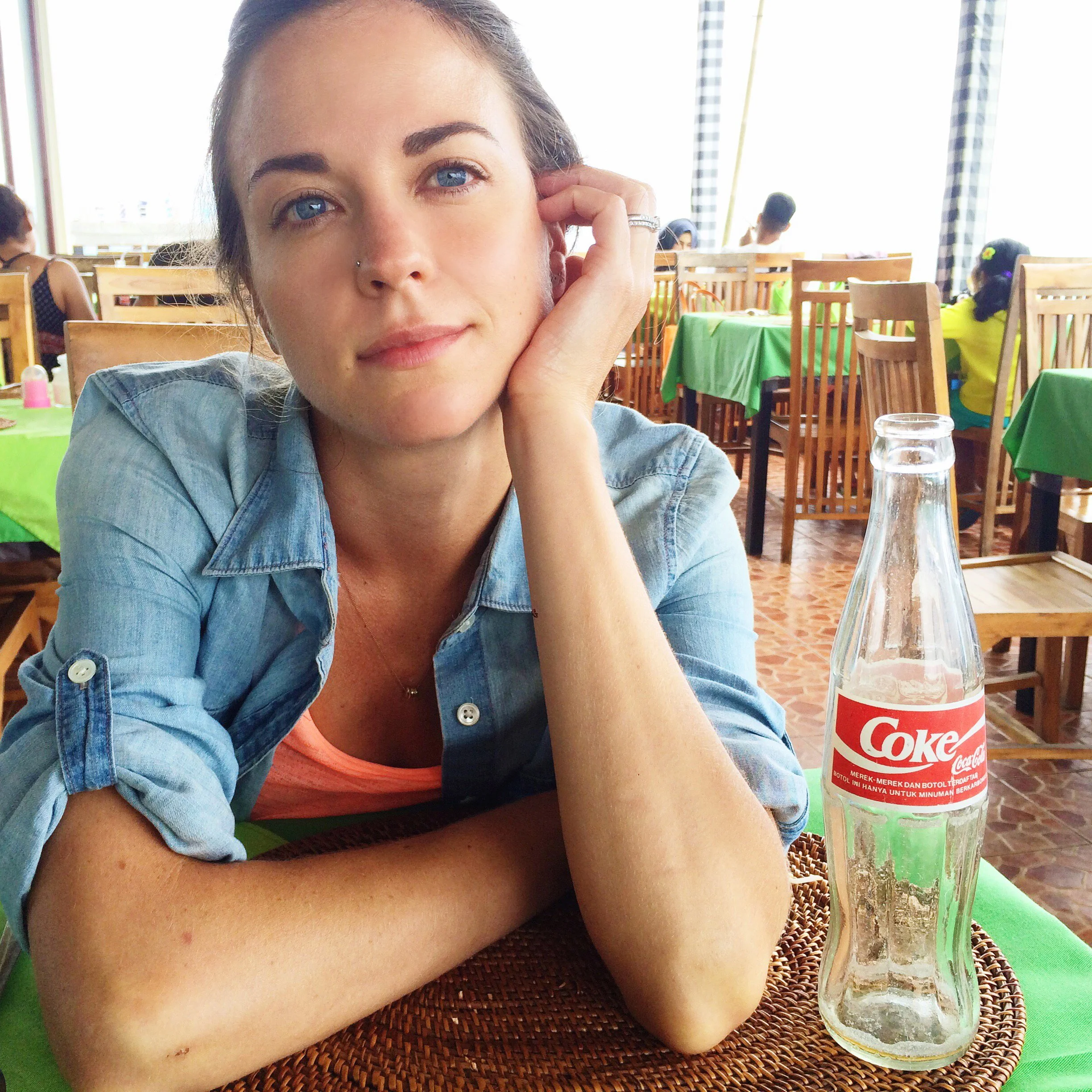 A woman with blue eyes and fair skin sitting at a table in a restaurant, resting her head on her right hand, with a glass bottle of Diet Coke in front of her, during daytime.