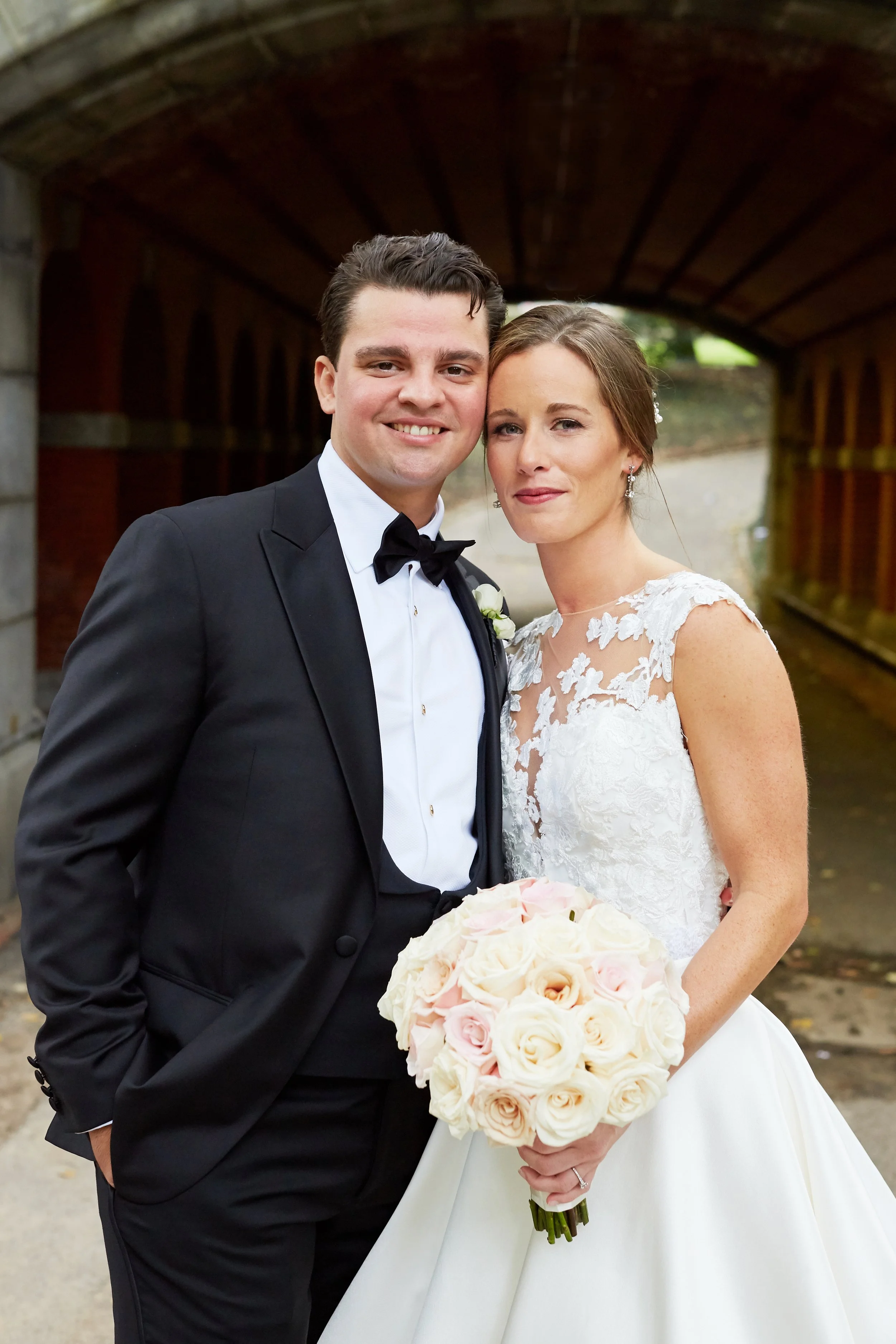 A newlywed couple dressed in wedding attire standing under a brick archway, with the bride holding a bouquet of white and blush pink roses.