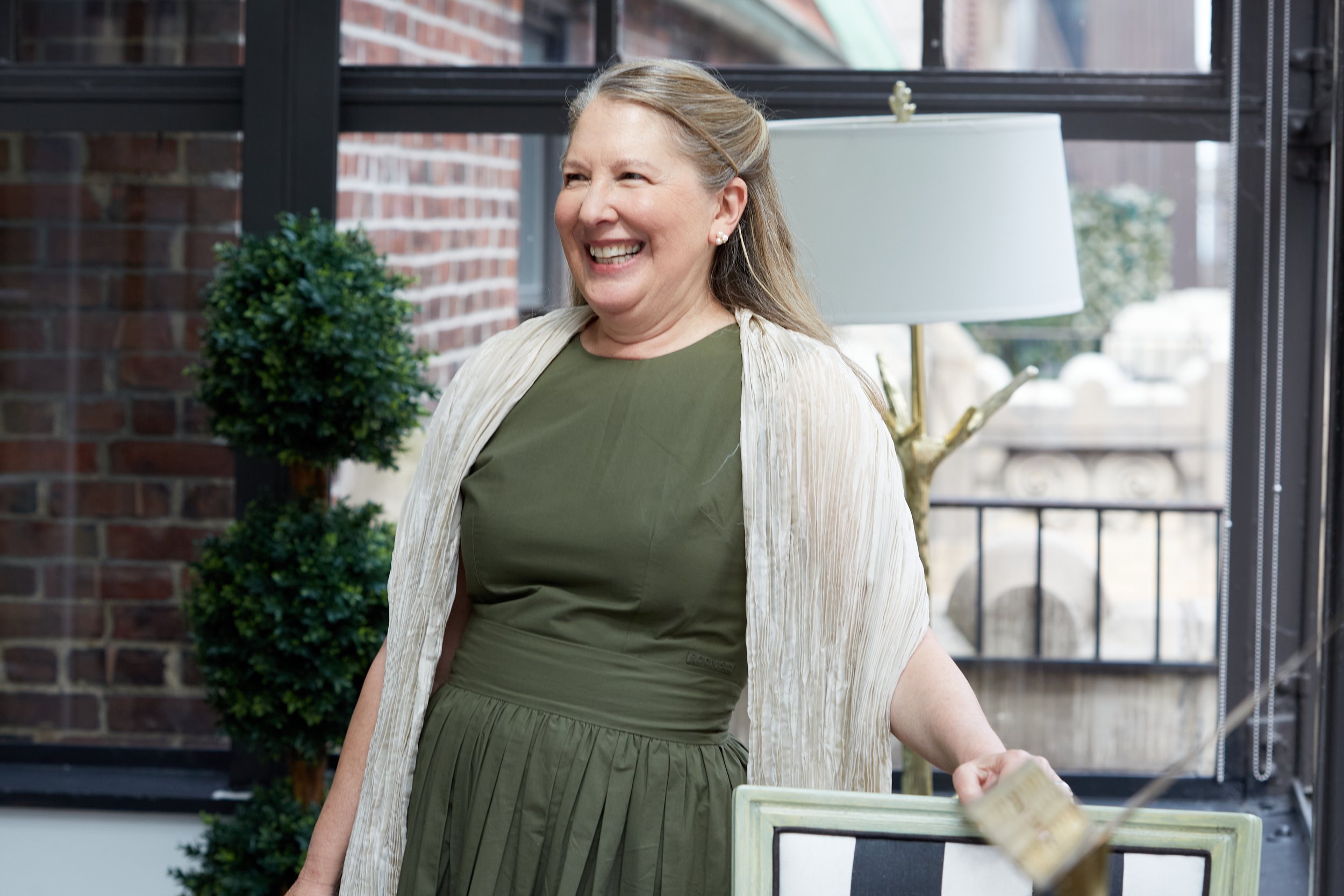 A smiling woman with blonde hair wearing a green dress and a light scarf, standing indoors near a window with plants and a lamp in the background.