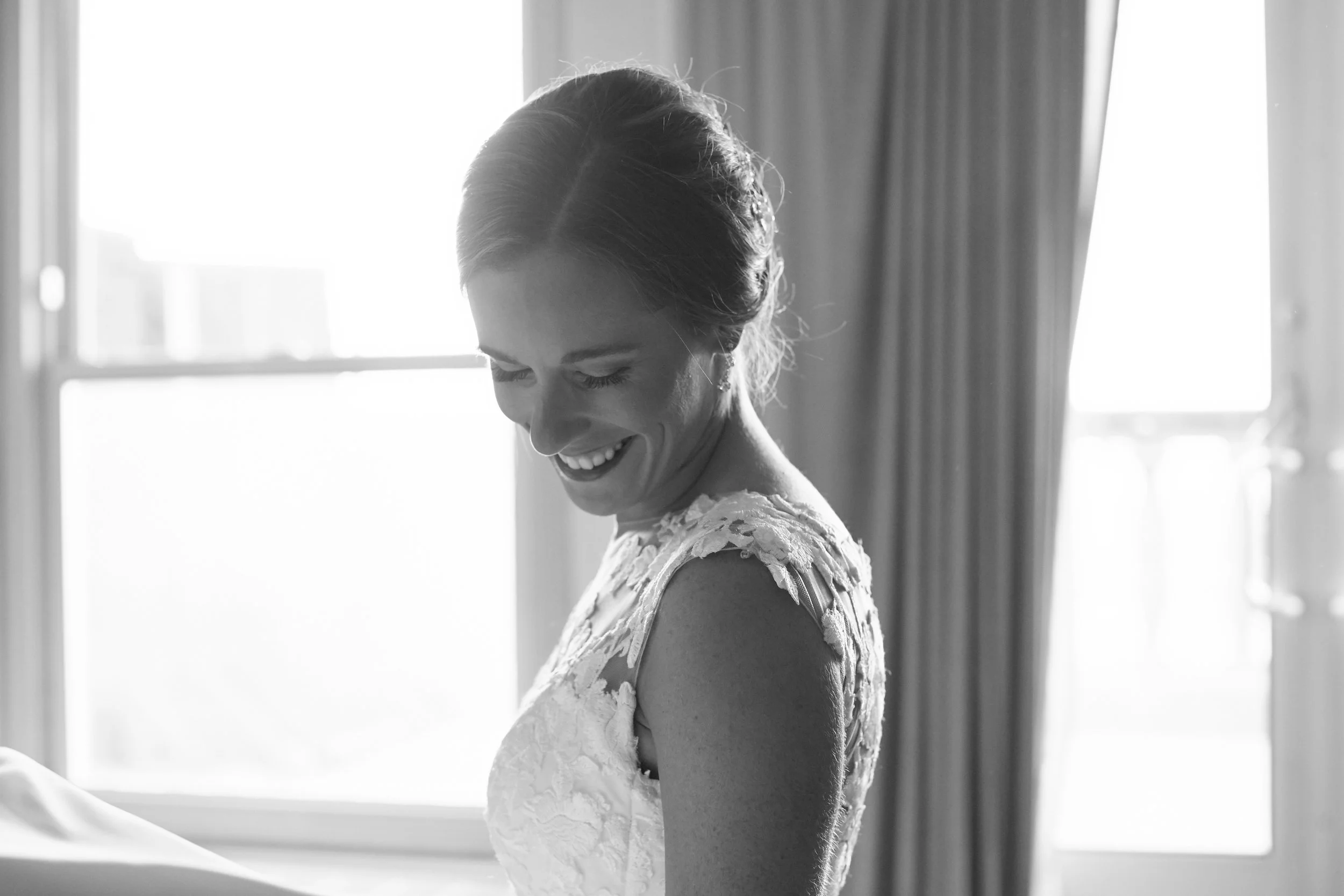 Black and white photo of a smiling woman in a lace dress, looking down, standing indoors near a window and curtains.
