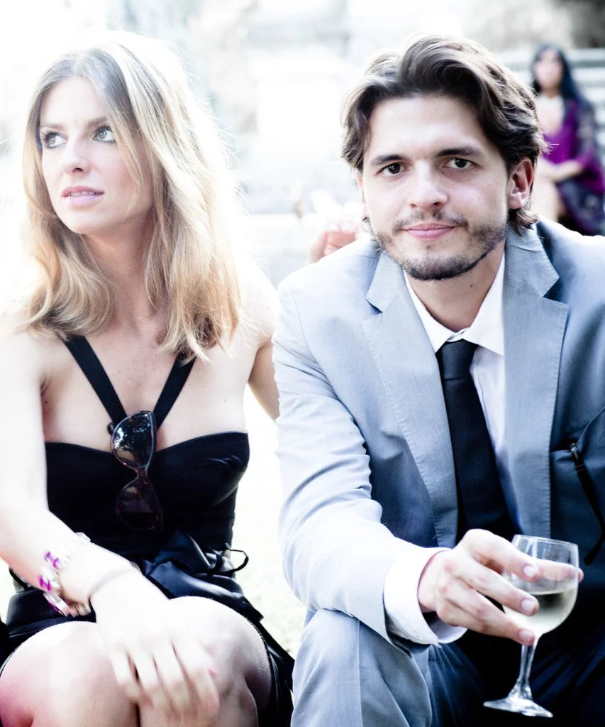A woman in a black dress and a man in a gray suit with a black tie sitting outdoors, with the man holding a glass of white wine.