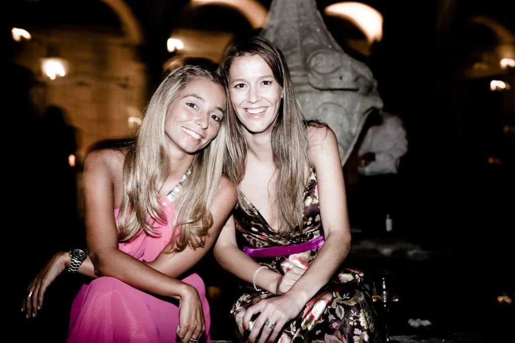 Two young women smiling and sitting close together at a nighttime event, in an indoor setting with dim lighting and a stone sculpture or fountain in the background.