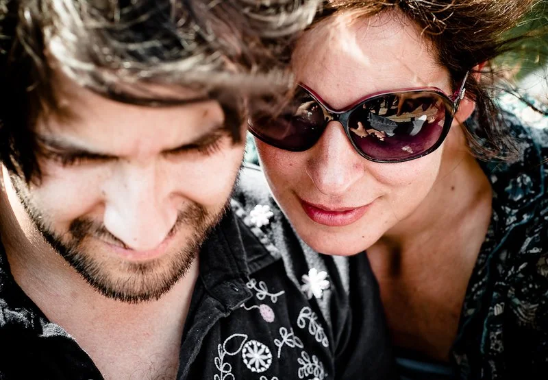 Close-up of a smiling man with a beard and a woman wearing sunglasses, both looking downward, outdoors.