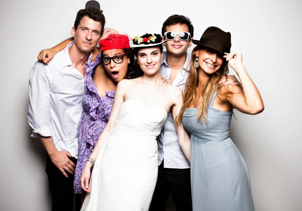 Five young adults standing together, dressed up and wearing playful accessories like hats and sunglasses, posing in front of a plain white background.