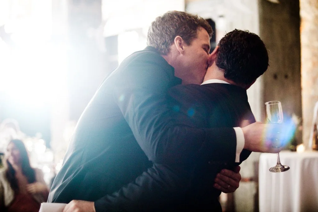 Two men, dressed in tuxedos, embracing and kissing at a celebration, with one holding a glass of champagne.