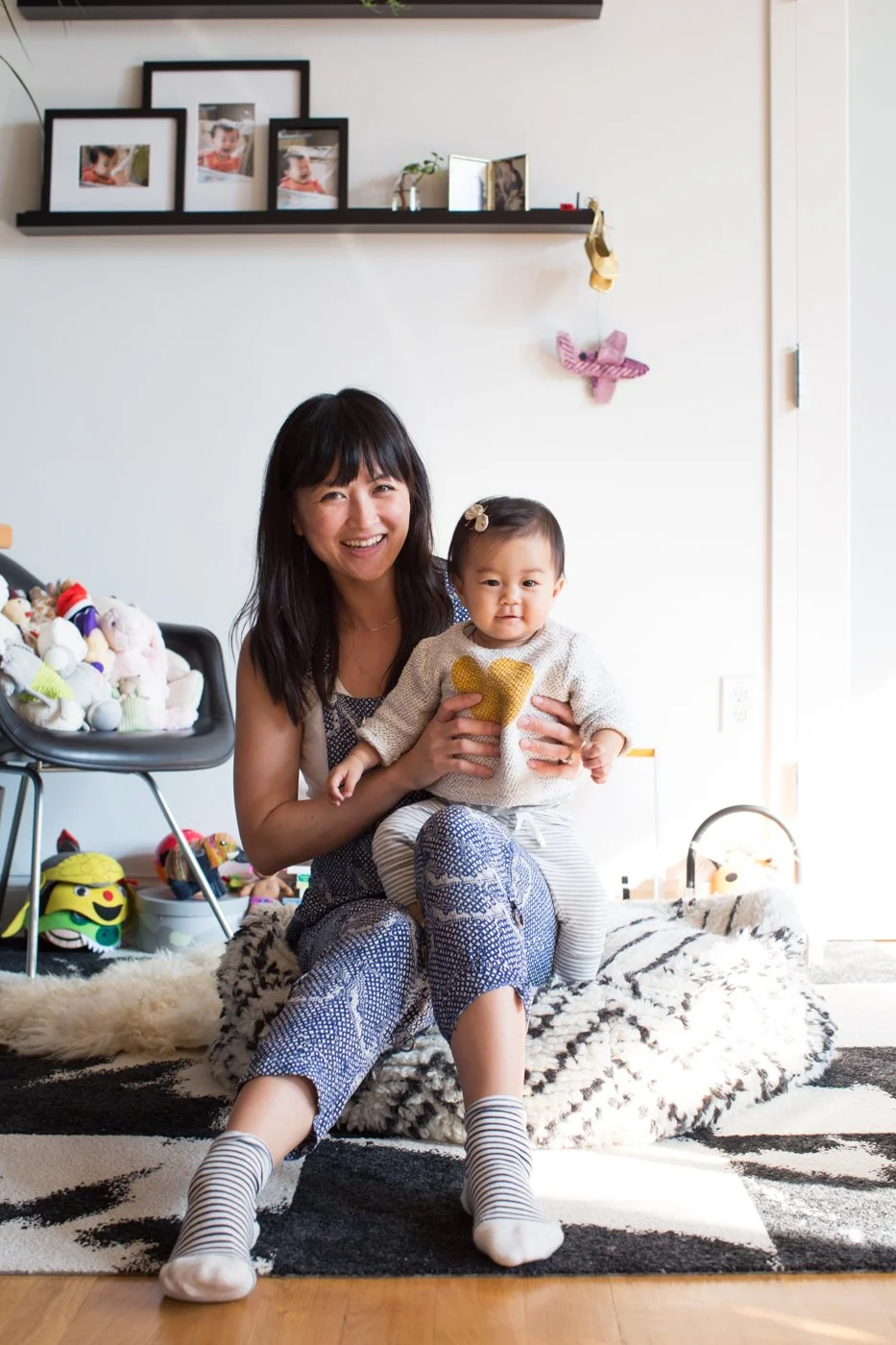 A woman sitting on a black and white rug holds a young girl with a beige sweater with a gold heart on it. They are smiling inside a brightly lit living room with toys on a chair, picture frames on a wall shelf, and a plush animal rug.