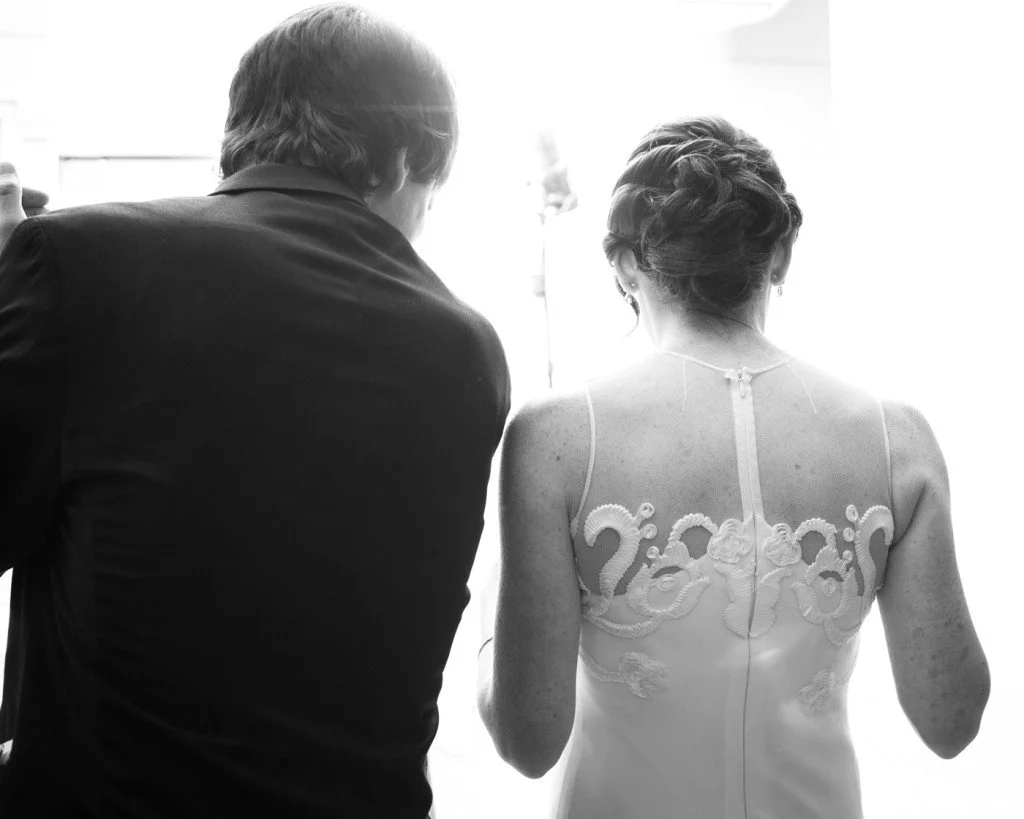 Back view of a woman in a wedding dress and a man in a dark suit, sitting together.